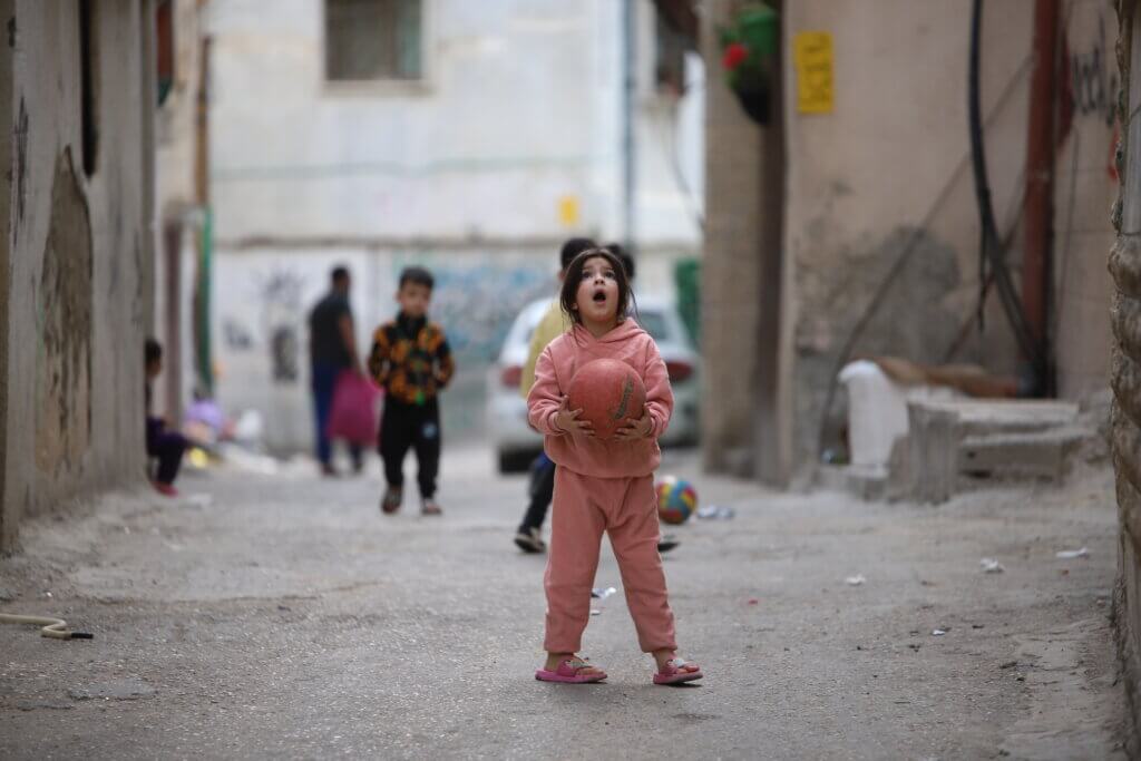 A Palestinian girl looks up in surprise while she plays with a ball in the middle of the street in a neighborhood in the Aida refugee camp, in the occupied West Bank city of Bethlehem. (Malik Hamamra/Mondoweiss) May 2023