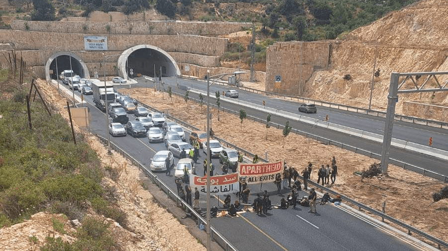 A photo from a distance of activists shutting down a main highway connecting West Bank settlements to Jerusalem. The activists are seen sitting in one of the highway lanes with a blocking a line of cars, with two large unfurled banners. One banner reads, 