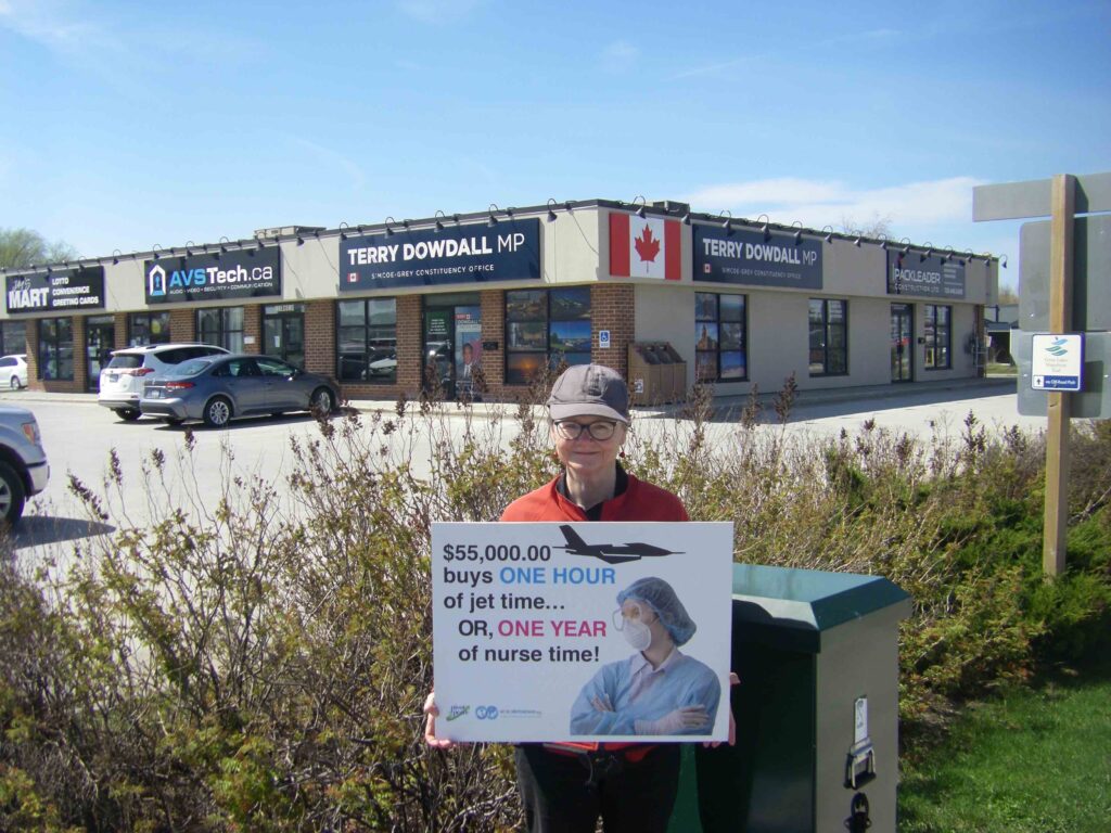 WBW Chapter member Gillian stands outside MP office with a sign reading $55,000 buys ONE HOUR of jet rime.. or ONE YEAR of nurse time!